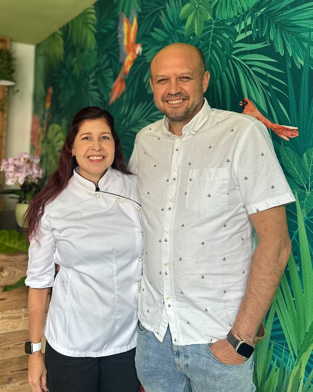 La chocolatera (izquierda), con chaqueta blanca de cocina, y su marido, con camisa blanca, propietarios de la Chocolaterie Tovar, en la tienda, delante de una pared empapelada con motivos de selva