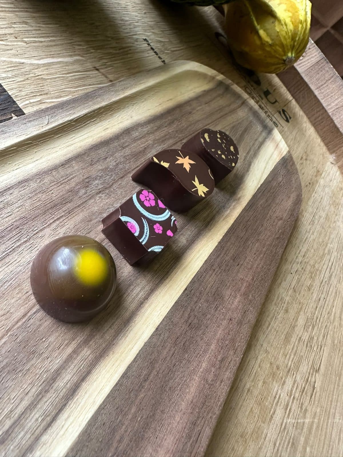 Cuatro bombones con decoraciones de colores sobre una tabla de madera y, al lado, una mazorca de cacao